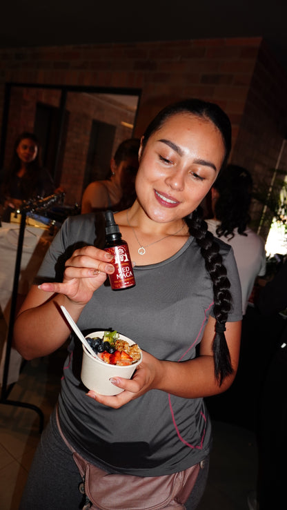 Person holding a Red Maca drink bottle while smiling in a warm indoor setting.